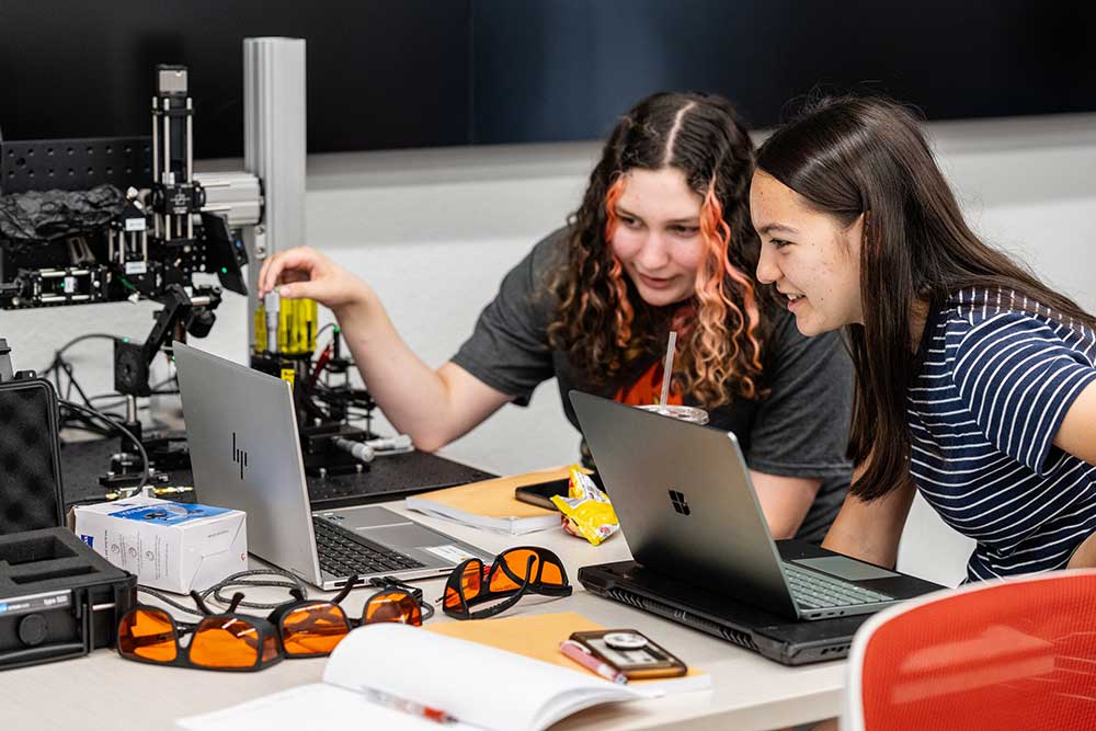 Table with shaded glasses, science instruments and computers. Two girls working together looking at computer screen.